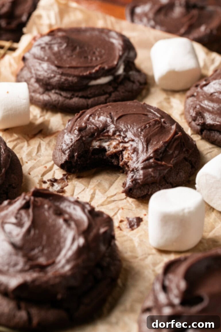 A delicious chocolate marshmallow cookie with a bite taken out, revealing its rich chocolate crumb and gooey marshmallow center, surrounded by other frosted cookies on parchment paper, highlighting its tempting quality.
