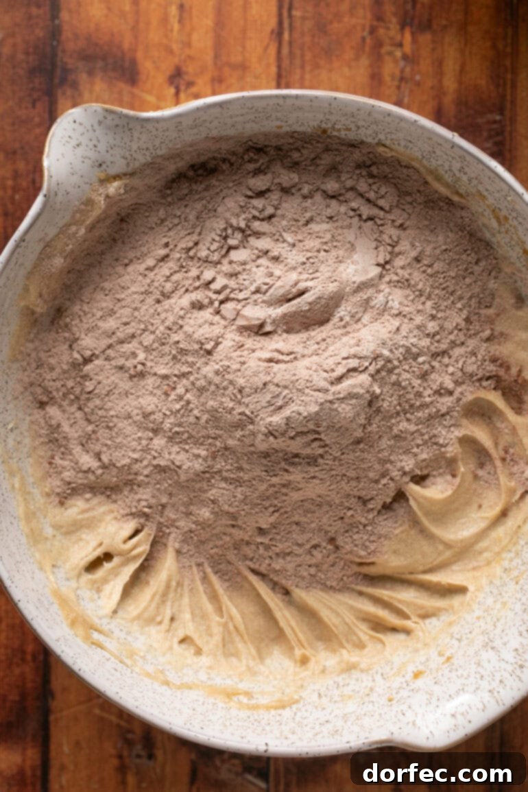 The dry cookie ingredients being gradually added to the wet mixture, showing the dough starting to come together into a rich, dark chocolate color, ready for final mixing.