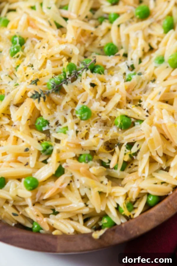 A close-up photo of a wooden bowl filled with creamy lemon orzo pasta, mixed with bright green peas, delicate shaved Parmesan cheese, and a sprinkle of freshly ground black pepper.