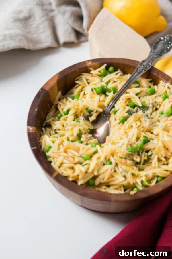 A silver spoon rests within a wooden bowl brimming with quick and easy lemon orzo pasta, accompanied by Parmesan cheese and peas, all presented on a pristine white table.