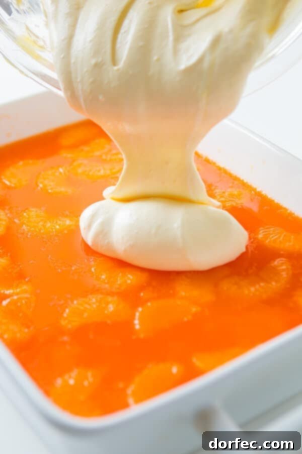 Sunshine Citrus Gelatin Delight 2 Someone pouring a smooth, white cool whip topping evenly over a pan of set orange jello salad in a white baking dish.
