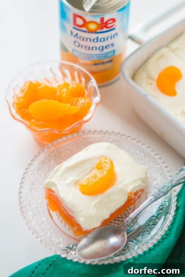 Lemon Mandarin Orange Jello Salad Serving A serving of lemon mandarin orange jello salad on a clear glass plate with a spoon, showcasing the distinct layers of orange jello and creamy lemon topping.