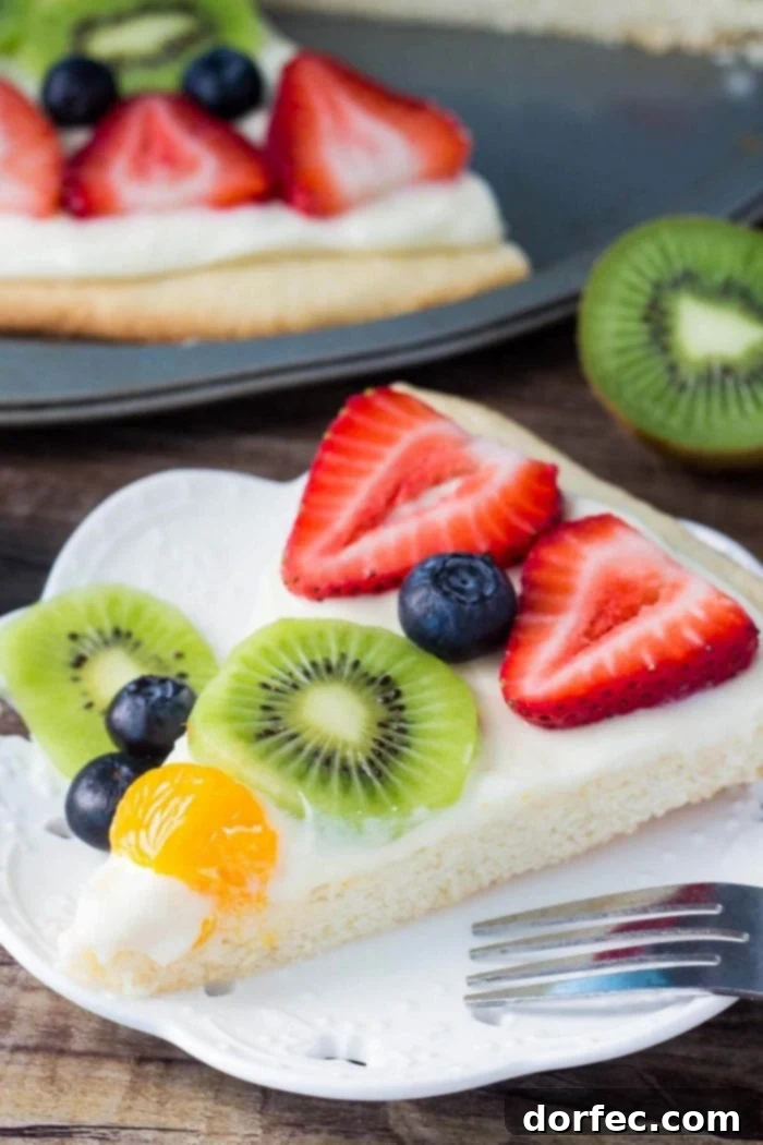 A perfectly sliced piece of sugar cookie fruit pizza on a white plate with a fork, showcasing its soft cookie base, creamy frosting, and fresh fruit toppings.