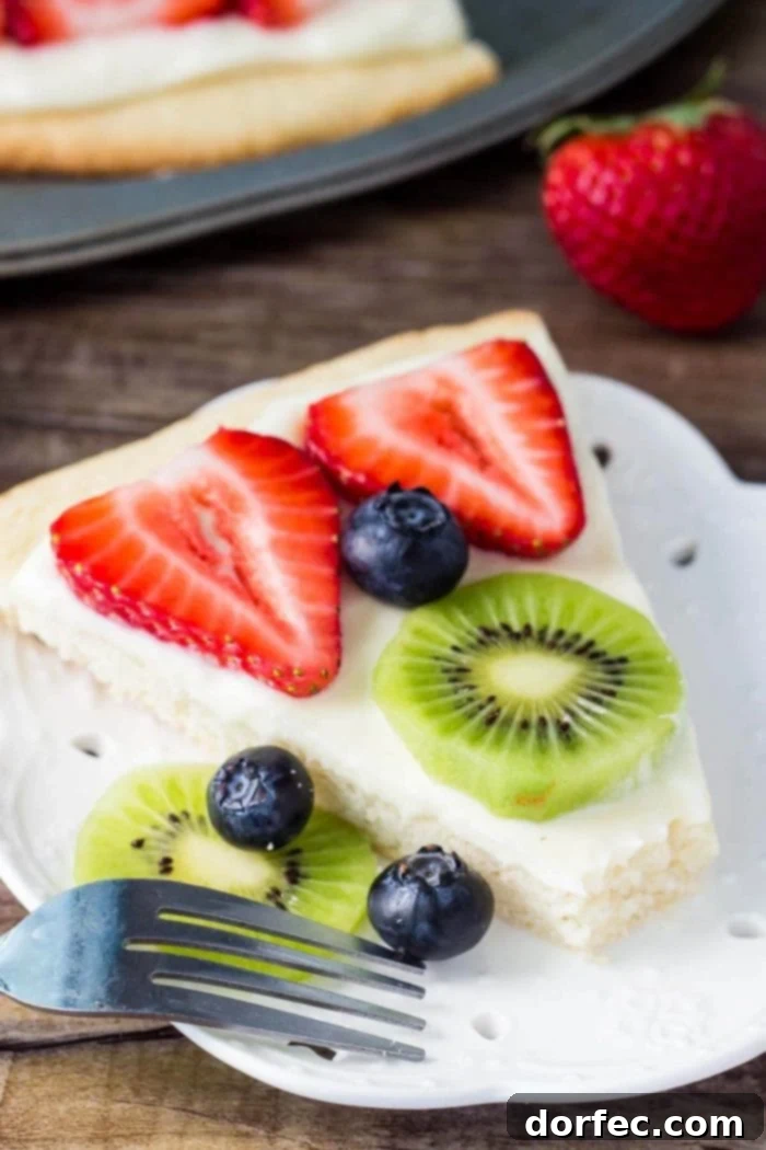 Close-up shot of a single slice of sugar cookie fruit pizza, perfectly portioned on a white plate, highlighting the delicious layers of cookie, frosting, and fruit.