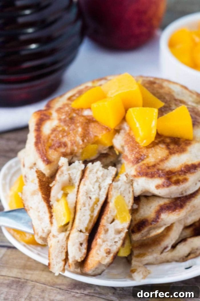 Fluffy Peach Pancakes Cut and Ready to Serve A stack of golden brown peach pancakes on a white plate, cut into, revealing the fluffy interior and juicy peach pieces. The image highlights the inviting texture and deliciousness of this breakfast.