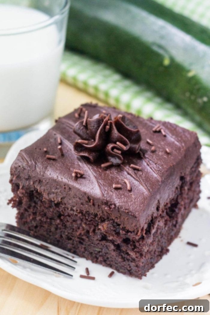 A perfectly frosted slice of double chocolate zucchini cake on a white plate, accompanied by a refreshing glass of milk, highlighting its rich, moist texture.