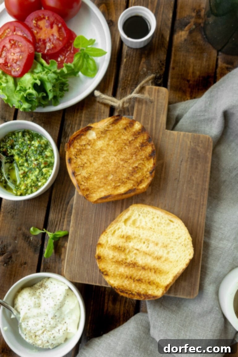 a photo of an open brioche bun laying face up on a wooden cutting board you can see the grill marks on the bun and there is a small bowl of pesto sitting next to it and a plate of lettuce and tomatoes.
