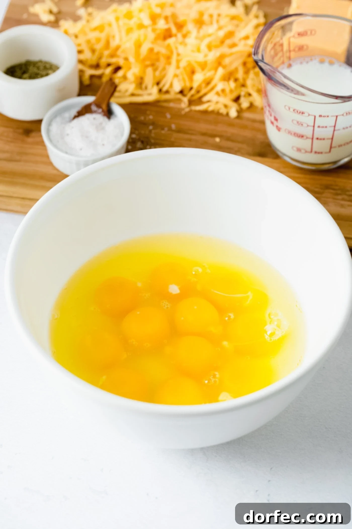 Essential Scrambled Egg Ingredients A pristine white bowl filled with raw eggs, ready for scrambling. In the background, ingredients like salt, shredded cheese, and a carton of milk are artfully arranged.