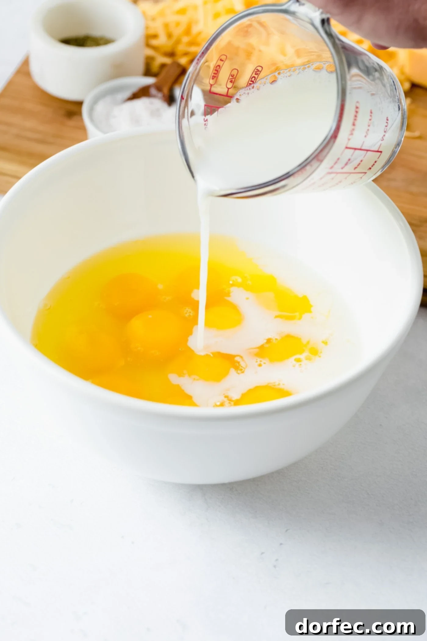 Mixing Scrambled Egg Ingredients A bowl of raw eggs, with fresh milk being poured over them, creating a creamy mixture. Shredded cheese, salt, and pepper shakers are visible in the background, hinting at the seasonings to come.