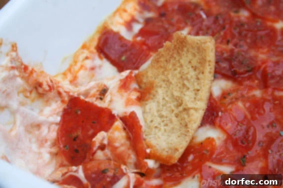 Ultimate Pepperoni Pizza Dip 3 a pita chip dunked into a baking dish of pepperoni pizza dip, showing the gooey cheese