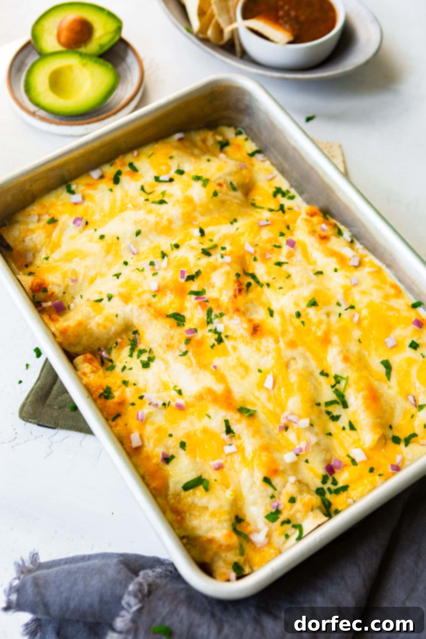 A photo of a 9x13 casserole dish full of saucy Honey Lime Chicken Enchiladas, garnished with melted cheese, diced red onions, and chopped cilantro, fresh from the oven.