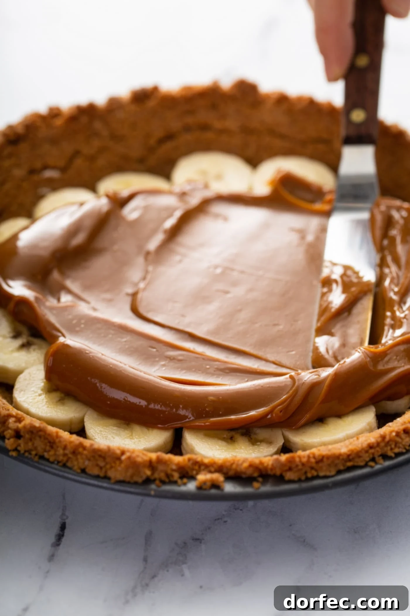 Decadent Dulce De Leche Banana Cream Pie 15 A graham cracker crust in a pie dish. There are sliced bananas in the bottom of the crust and dulce de leche is being spread over the top of the bananas.