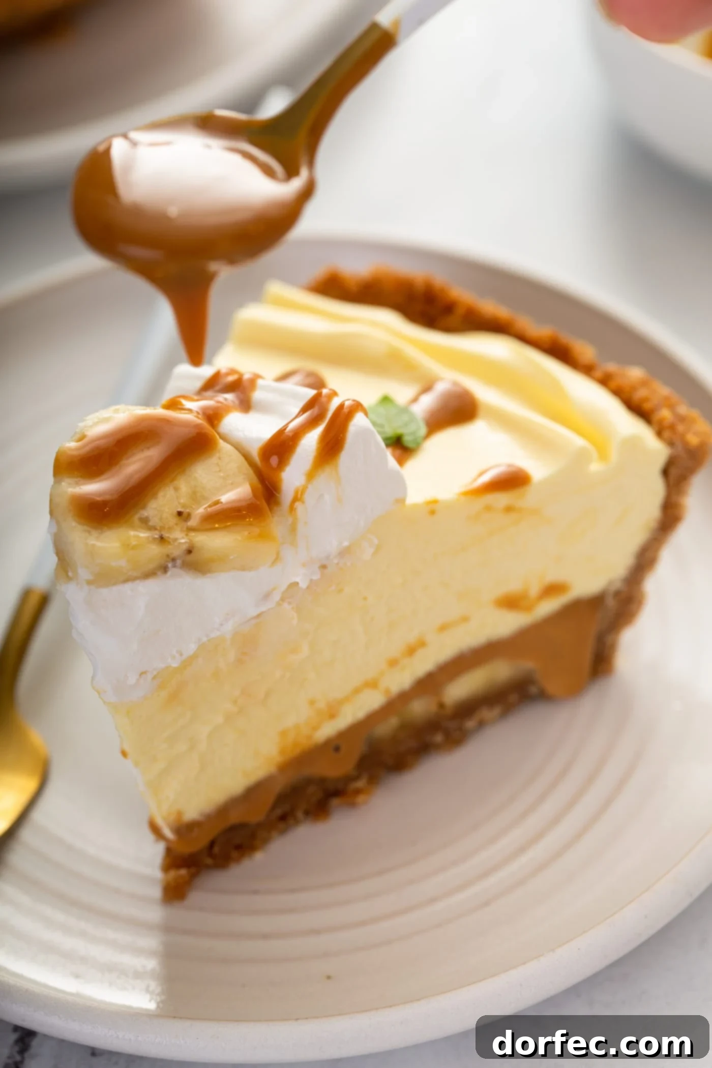 Decadent Dulce De Leche Banana Cream Pie 5 A slice of dulce de leche banana cream pie. The pie has a graham cracker crust and is a golden yellow color. There is a dollop of whipped cream on top and dulce de leche is being drizzled over the top of the whipped cream.