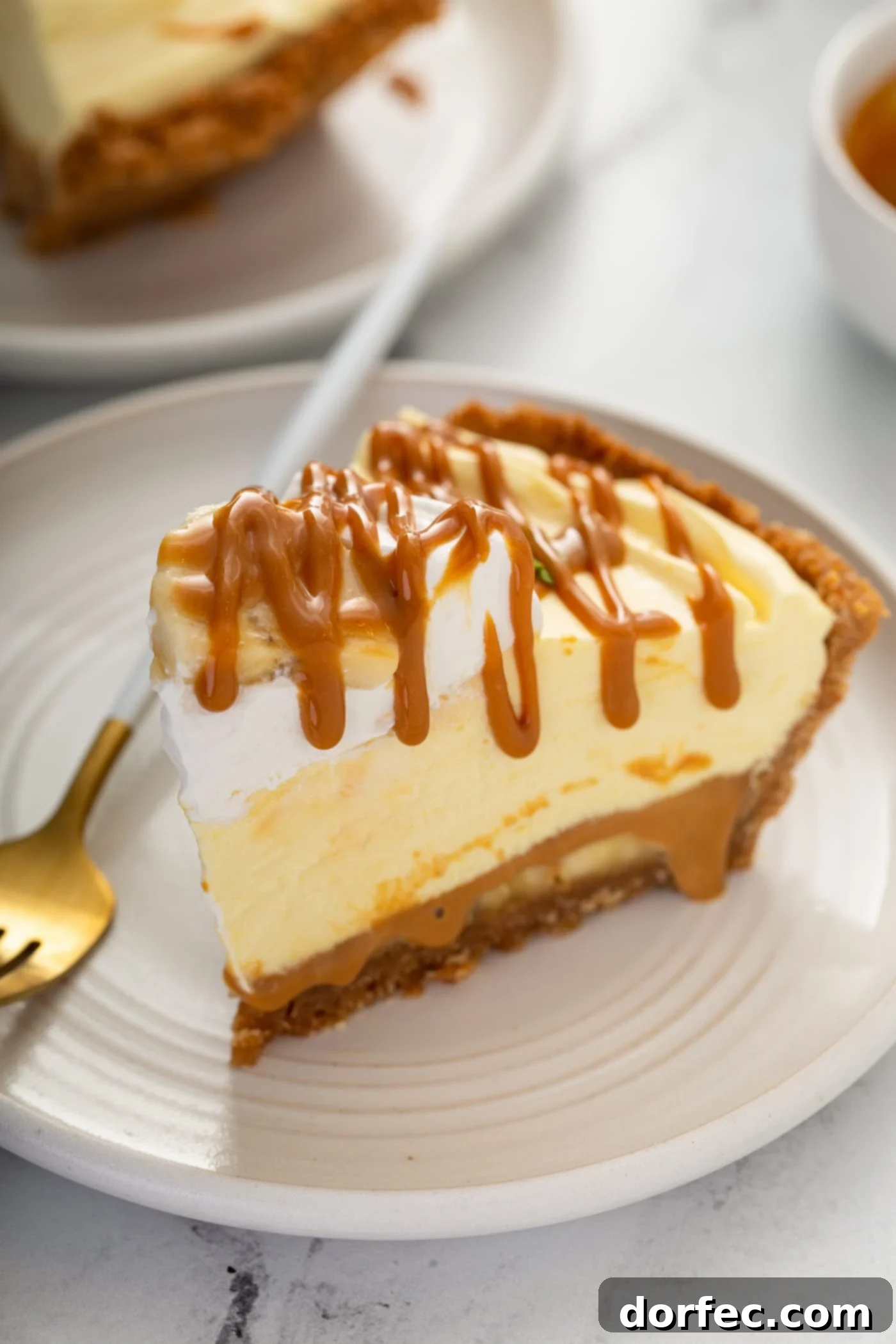 Decadent Dulce De Leche Banana Cream Pie 6 A slice of dulce de leche banana cream pie in a graham cracker crust. there is whipped cream on top and dulce de leche is drizzled over the top of the slice of pie. A fork rests on the plate next to the pie.