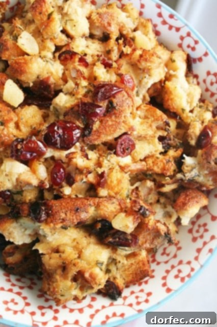 A serving spoon scoops a portion of Apple Cranberry Stuffing from a large baking dish, showing the moist interior with visible apple pieces and cranberries.