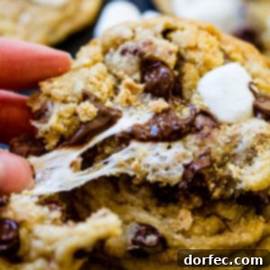 A close-up of a S'mores Chocolate Chip Cookie being broken in half, revealing the warm, stretching marshmallow and molten chocolate within.