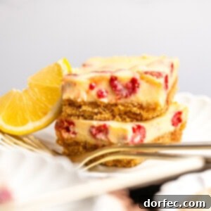 a photo of two creamy lemon raspberry bars with graham cracker crust stacked on each other on a small dessert plate with a lemon wedge next to them