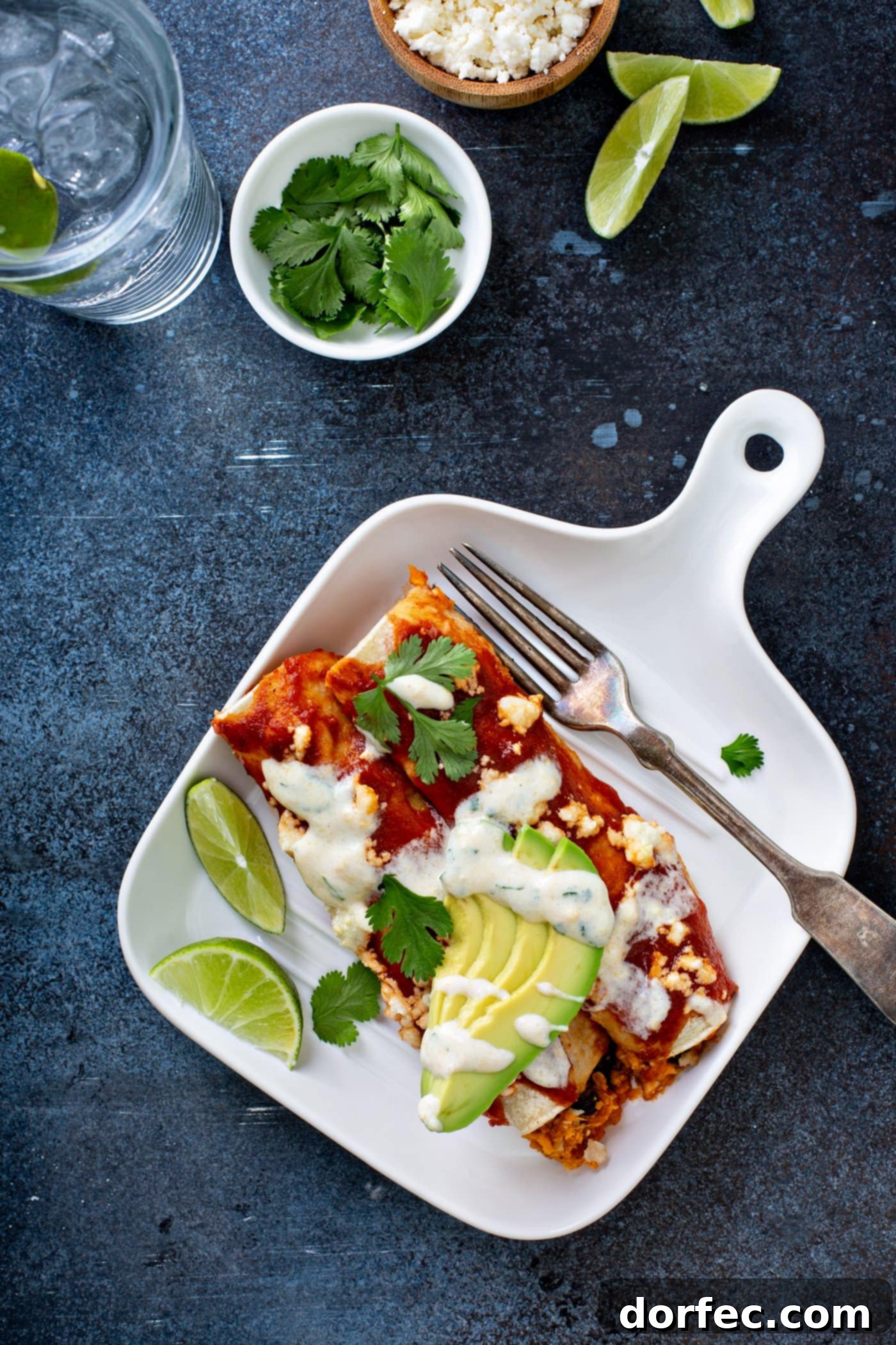 A dinner plate featuring sweet potato enchiladas, garnished with red enchilada sauce, fresh avocado slices, cilantro leaves, and a drizzle of homemade sweet corn crema. Lime wedges sit beside the dish, with a glass of water and fresh cilantro in the background, highlighting this vibrant vegetarian enchilada recipe.