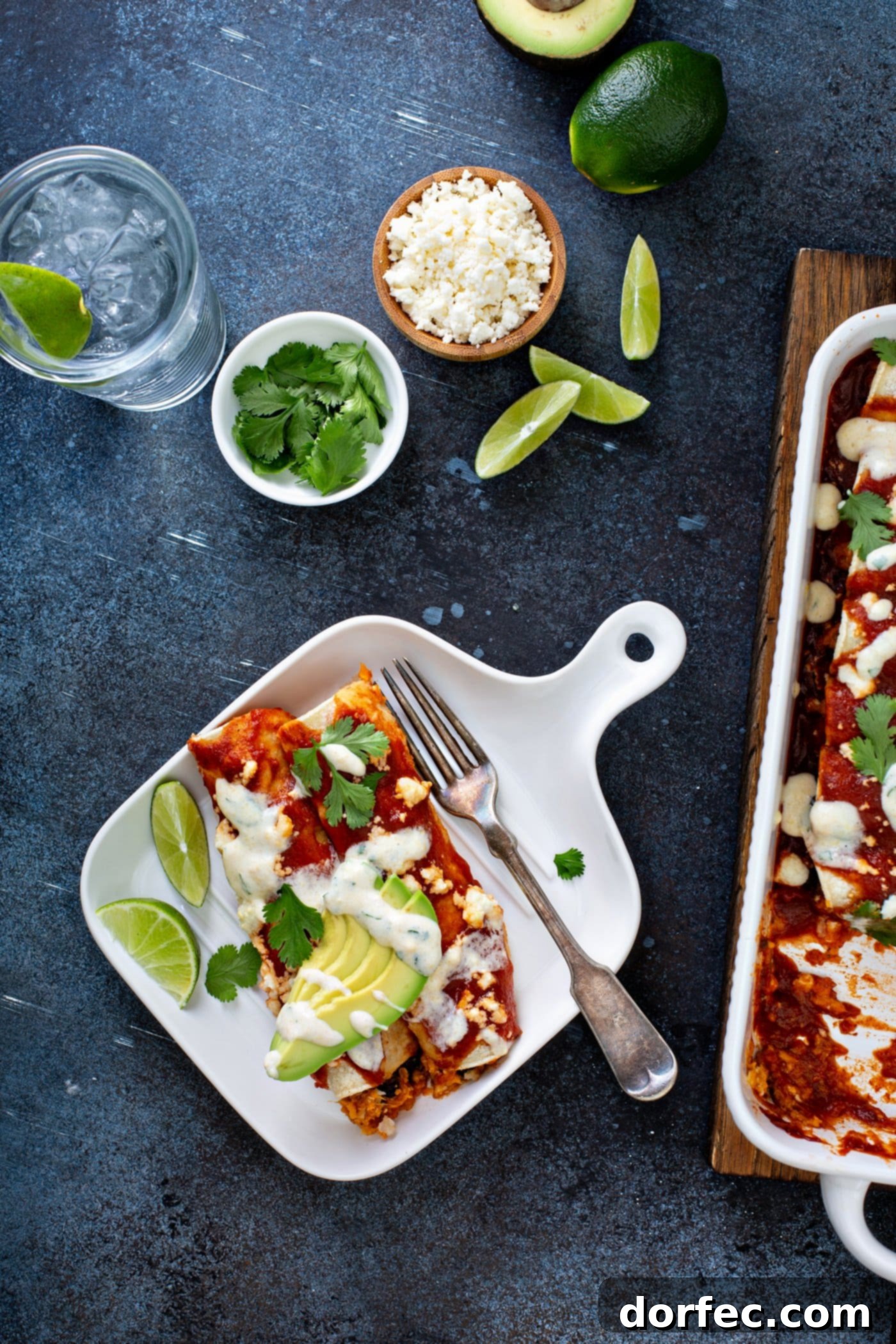 A serving of sweet potato enchiladas on a dinner plate, accompanied by a fork and lime wedges. The enchiladas are beautifully topped with red enchilada sauce, queso fresco, avocado slices, cilantro, and a drizzle of sweet corn crema. A baking dish of more enchiladas and various garnishes are in the background, showcasing a complete and vibrant vegetarian meal.