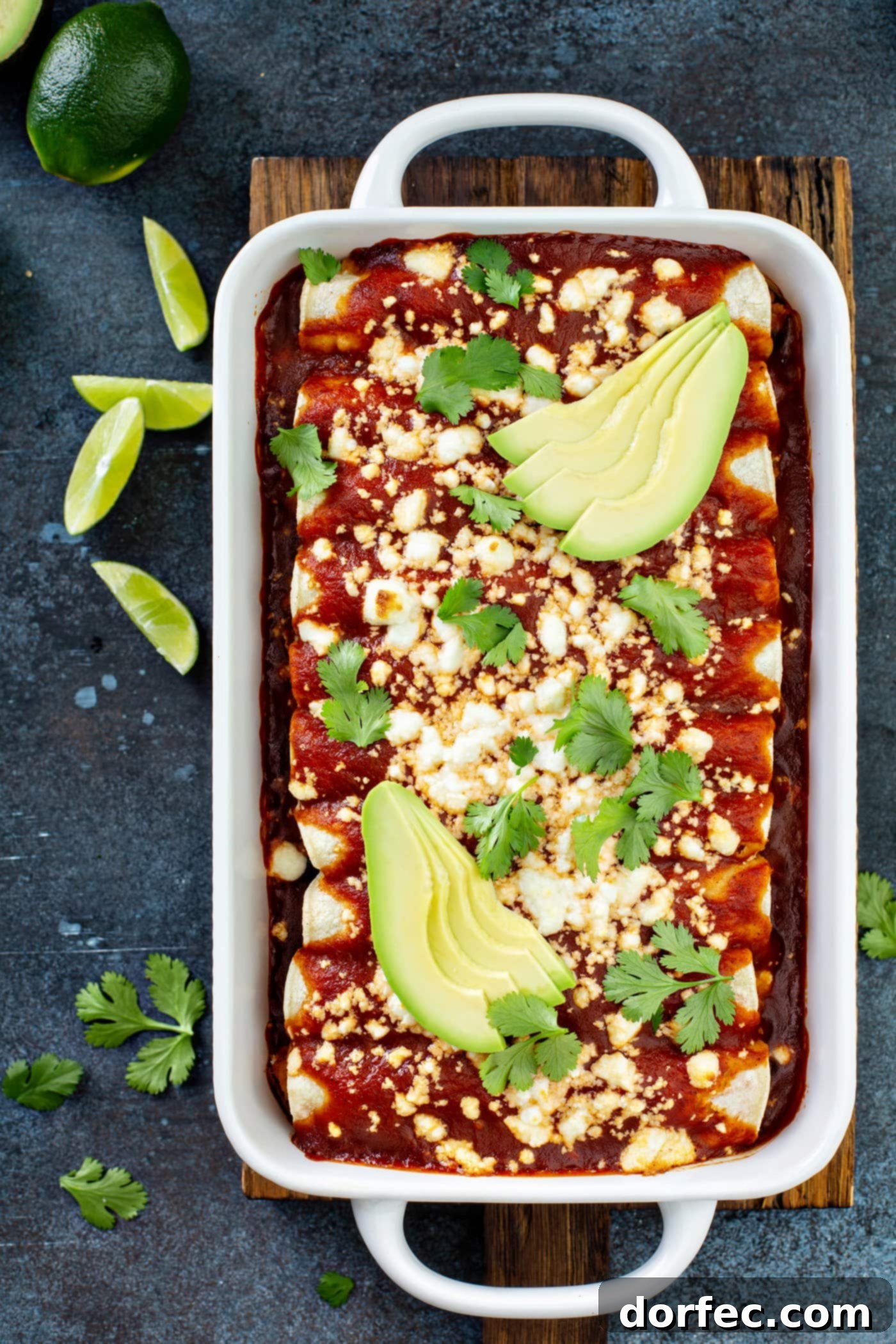A white casserole dish filled with baked sweet potato enchiladas, artfully arranged with a red enchilada sauce, queso fresco cheese, avocado slices, and fresh cilantro leaves. Lime wedges and cilantro leaves are scattered around the dish on a rustic table, highlighting this vibrant and healthy Mexican main dish.