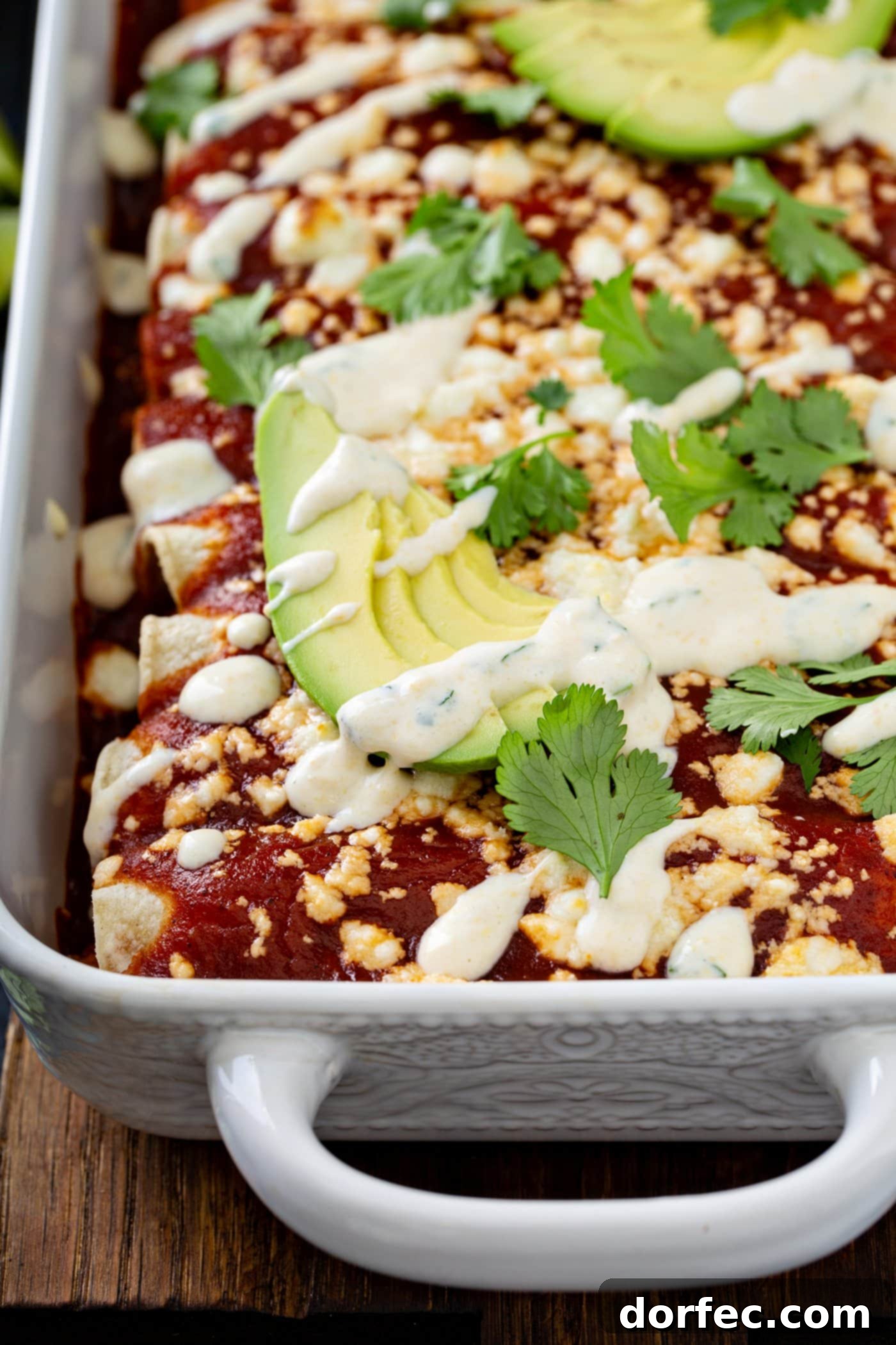 A close-up shot of baked sweet potato enchiladas in a baking dish, exquisitely topped with red enchilada sauce, crumbled queso fresco cheese, avocado slices, cilantro, and a delicate drizzle of sweet corn crema. This detailed view emphasizes the rich textures and vibrant garnishes of this healthy vegetarian Mexican dish.