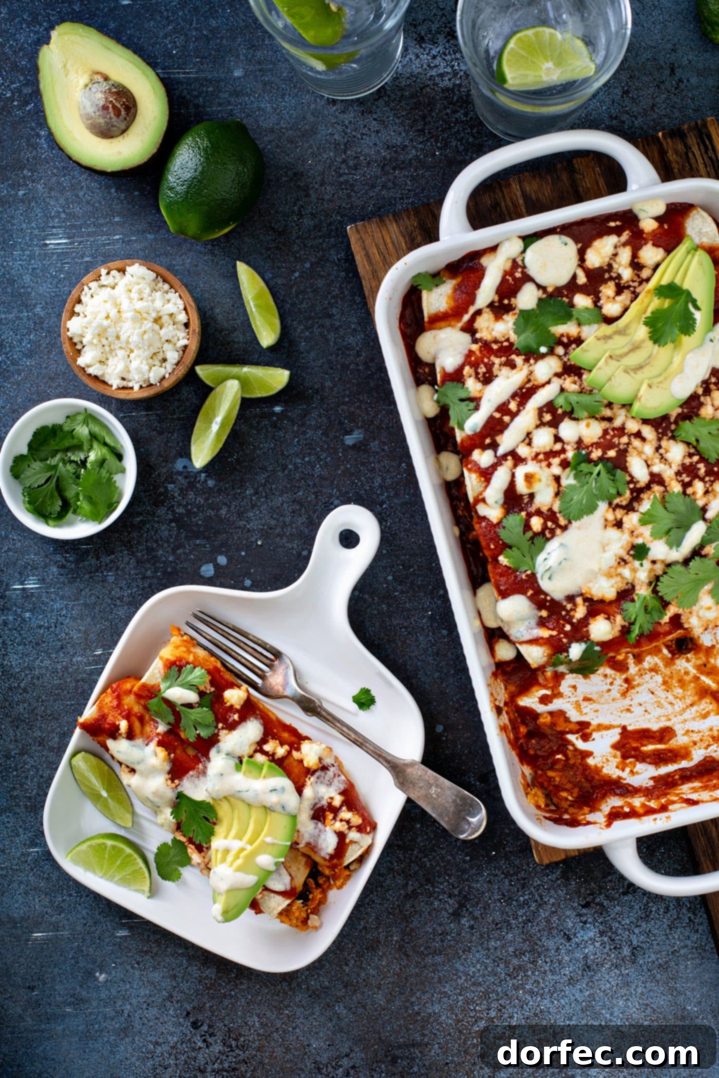 A dinner plate with a single serving of sweet potato enchiladas, garnished with red enchilada sauce, avocado slices, cilantro, and queso fresco cheese, all drizzled with sweet corn crema. A fork rests on the plate, with a casserole dish of remaining enchiladas and various garnishes in the background, showcasing a delicious and healthy vegetarian meal.