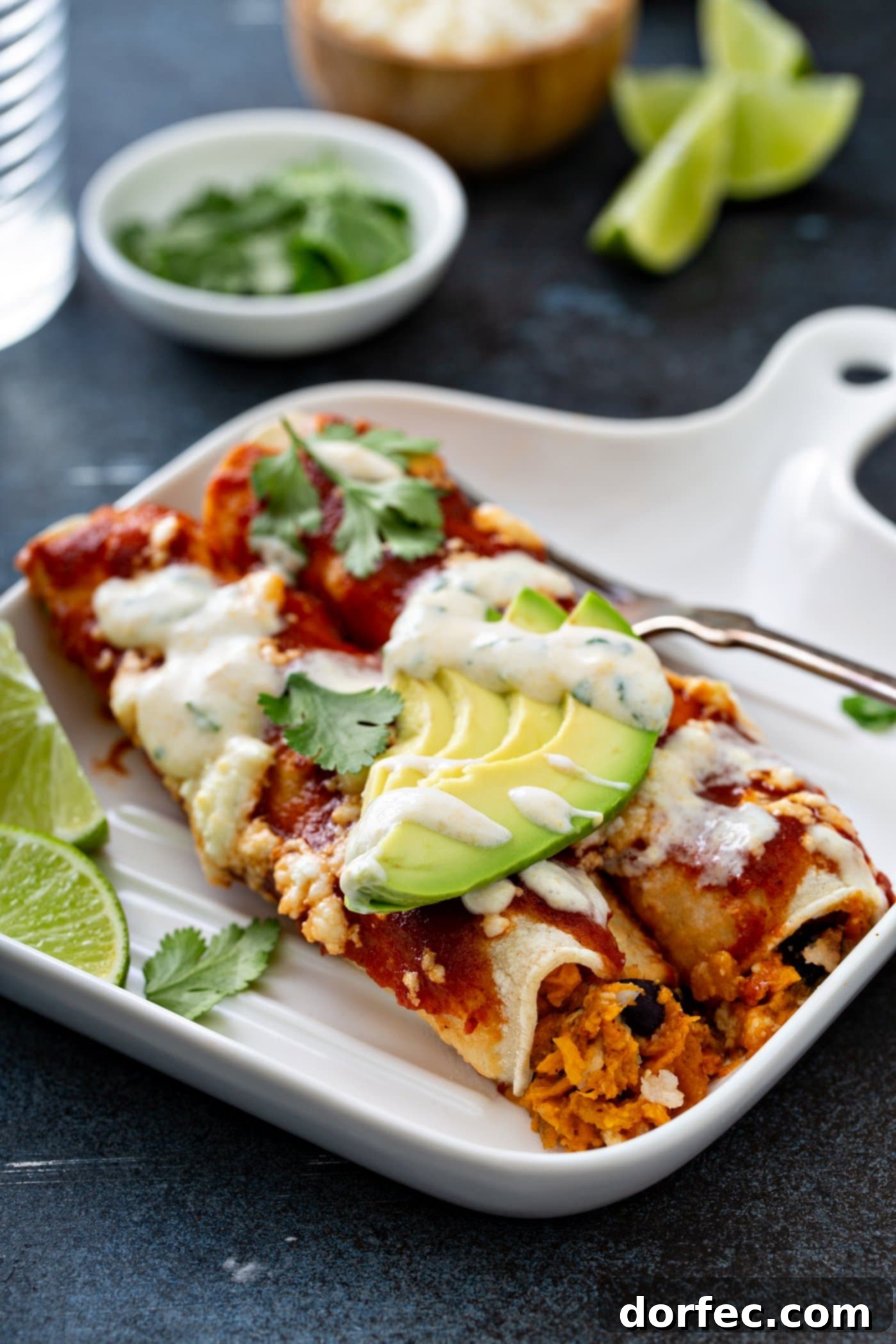 A dinner plate showcasing two sweet potato enchiladas, served with a fork and two lime wedges. The enchiladas are artfully garnished with red enchilada sauce, queso fresco cheese, avocado slices, cilantro leaves, and a delicate drizzle of sweet corn crema. This vibrant dish offers a healthy and delicious vegetarian Mexican meal.