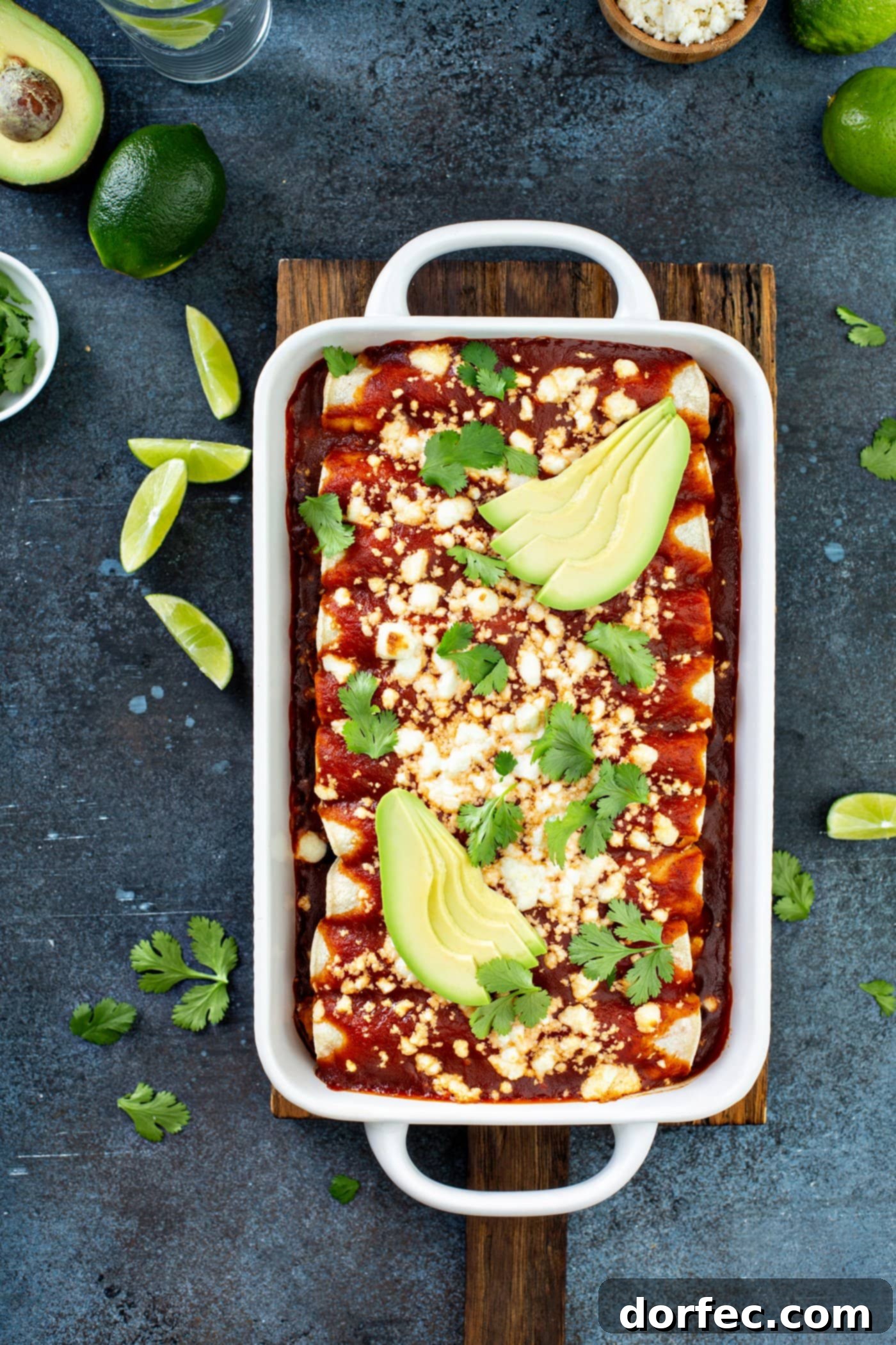 A white casserole dish filled with golden-brown sweet potato enchiladas, generously covered in red enchilada sauce and sprinkled with queso fresco cheese. Fresh avocado slices and cilantro leaves adorn the top, with lime wedges on the table, showcasing this delicious and healthy vegetarian Mexican main dish.