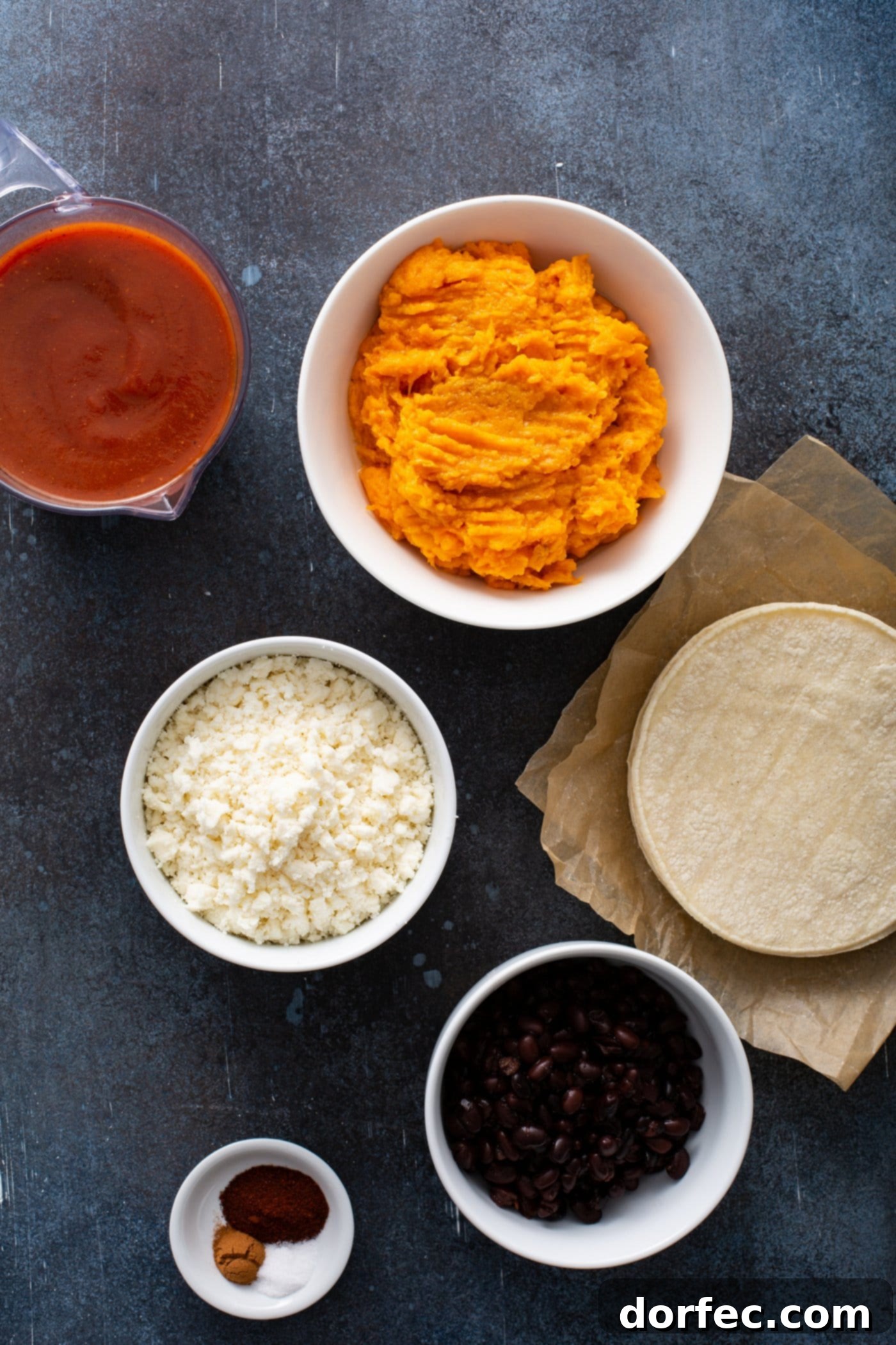 Small containers of mashed sweet potato, red enchilada sauce, crumbled queso fresco cheese, and corn tortillas, laid out as ingredients for sweet potato enchiladas. This image highlights the fresh, healthy components of the vegetarian enchilada recipe.