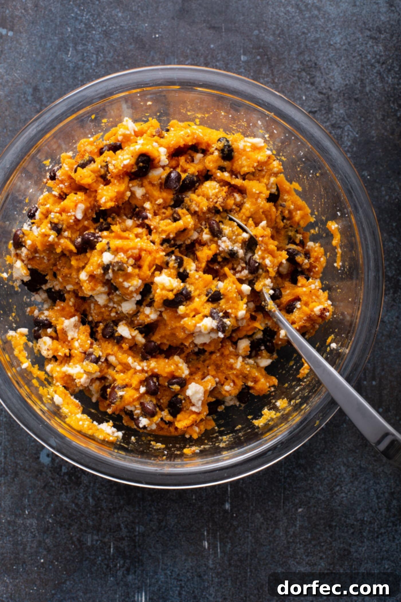 A glass bowl containing the sweet potato enchilada filling, a hearty mixture of mashed sweet potato, black beans, queso fresco, chili powder, and cinnamon, with a spoon resting in it. This image illustrates the rich, healthy filling for a vegetarian enchilada recipe.