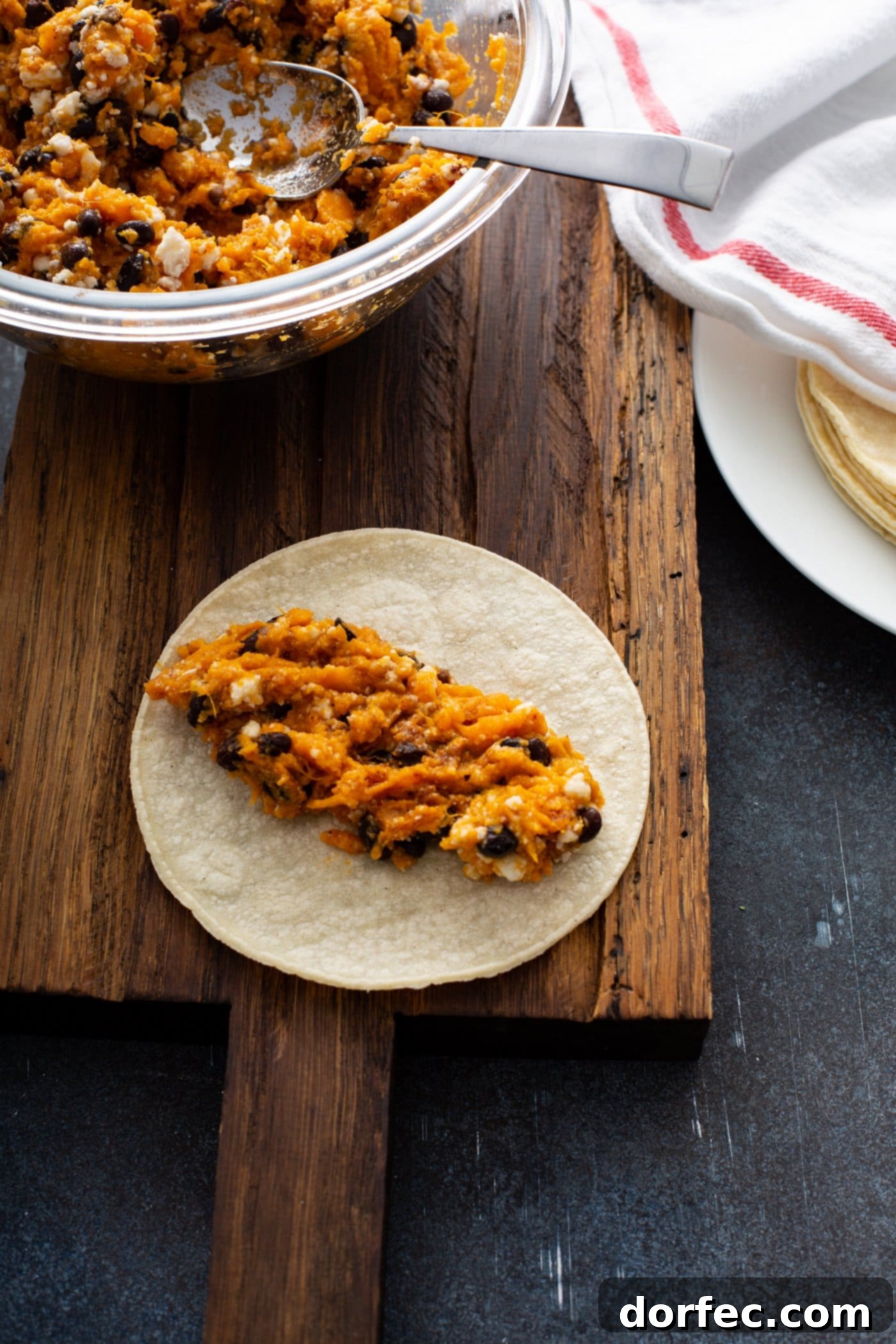 An open corn tortilla filled with sweet potato, black bean, and cheese mixture, ready to be rolled. In the background, a bowl of the delicious filling and a stack of corn tortillas covered with a kitchen towel await assembly. This step demonstrates the easy preparation of sweet potato enchiladas.