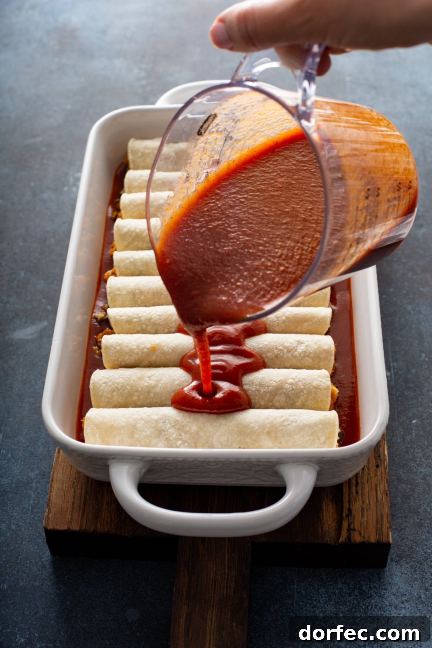 Unbaked sweet potato enchiladas arranged in a white baking dish, with red enchilada sauce being poured over them from a glass measuring cup. This step shows the preparation before baking for a rich and flavorful vegetarian enchilada dish.