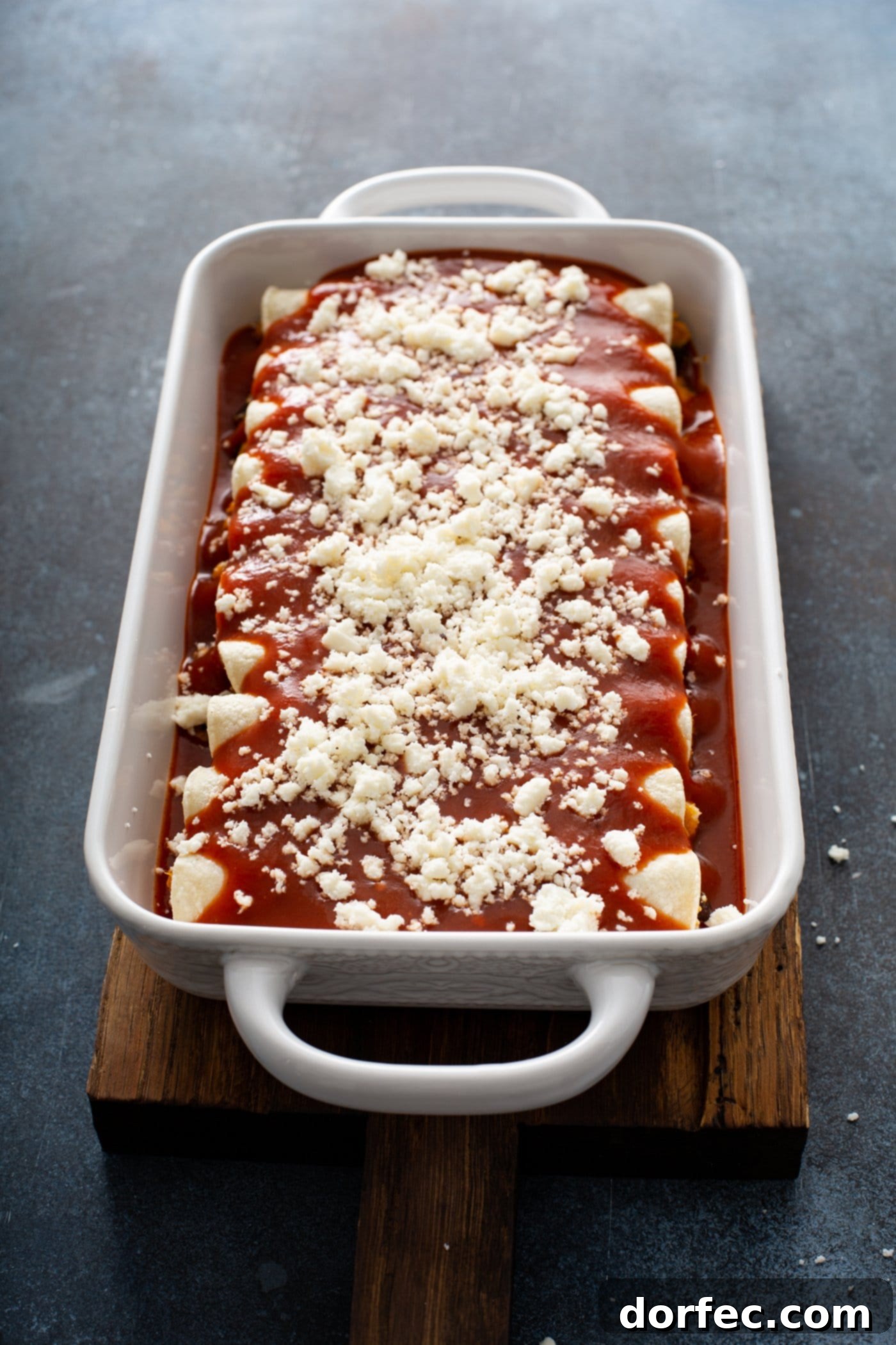 A casserole dish filled with unbaked sweet potato enchiladas, topped with vibrant red enchilada sauce and a generous sprinkle of queso fresco cheese. This image captures the dish just before it goes into the oven, promising a cheesy and hearty vegetarian meal.