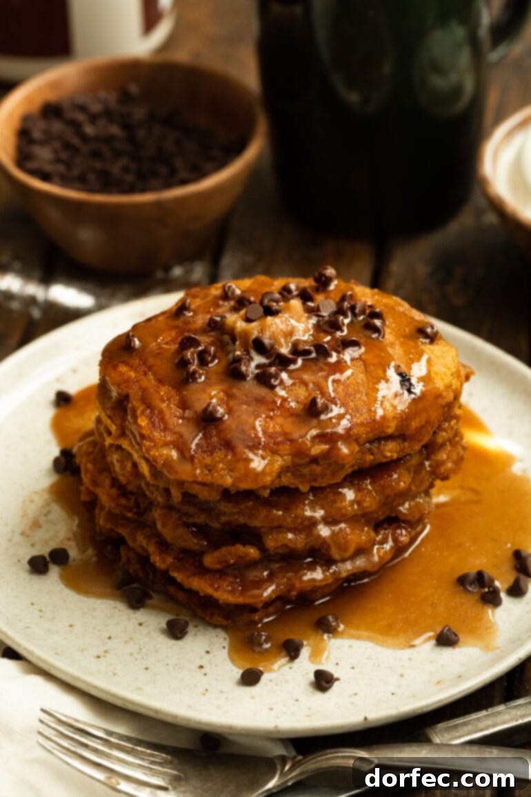 A stack of golden whole wheat pumpkin pancakes, lavishly coated in shimmering cinnamon caramel syrup and adorned with mini chocolate chips, set on a pristine white plate.