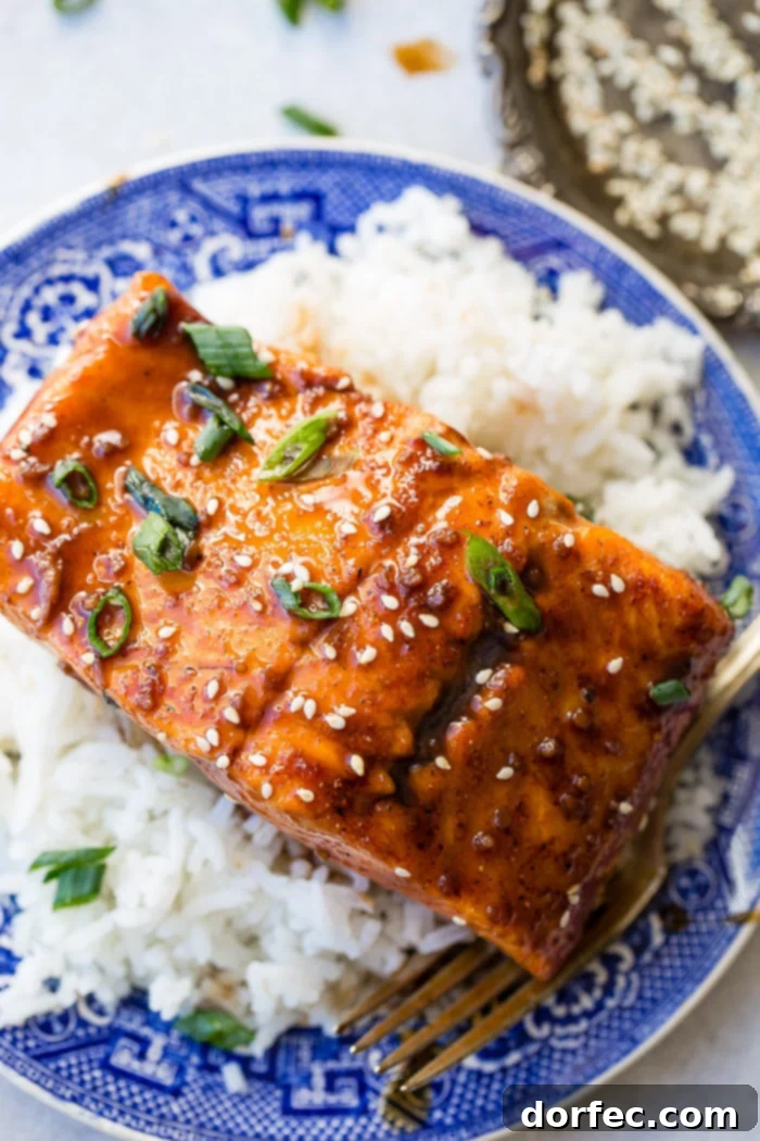Sweet & Sticky Asian Salmon Packs 3 A glazed fillet of salmon with green onions and sesame seeds, served on a bed of white rice on an ornate blue Asian-printed china dish.