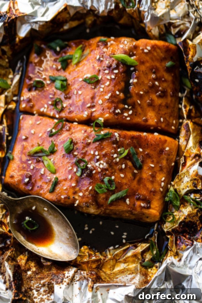 Sweet & Sticky Asian Salmon Packs 4 Two fillets of Asian Sticky Salmon in foil, garnished with green onions and sesame seeds, alongside a vintage spoon with sauce.