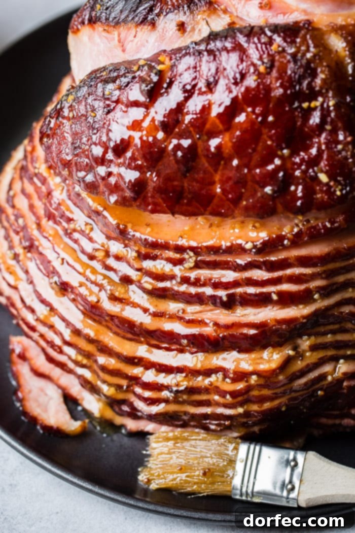 A close-up view of a beautifully glazed spiral ham with a golden-brown crust, basted with a rich honey mustard sauce and garnished on a sleek black platter. A basting brush rests alongside, indicating its fresh application.