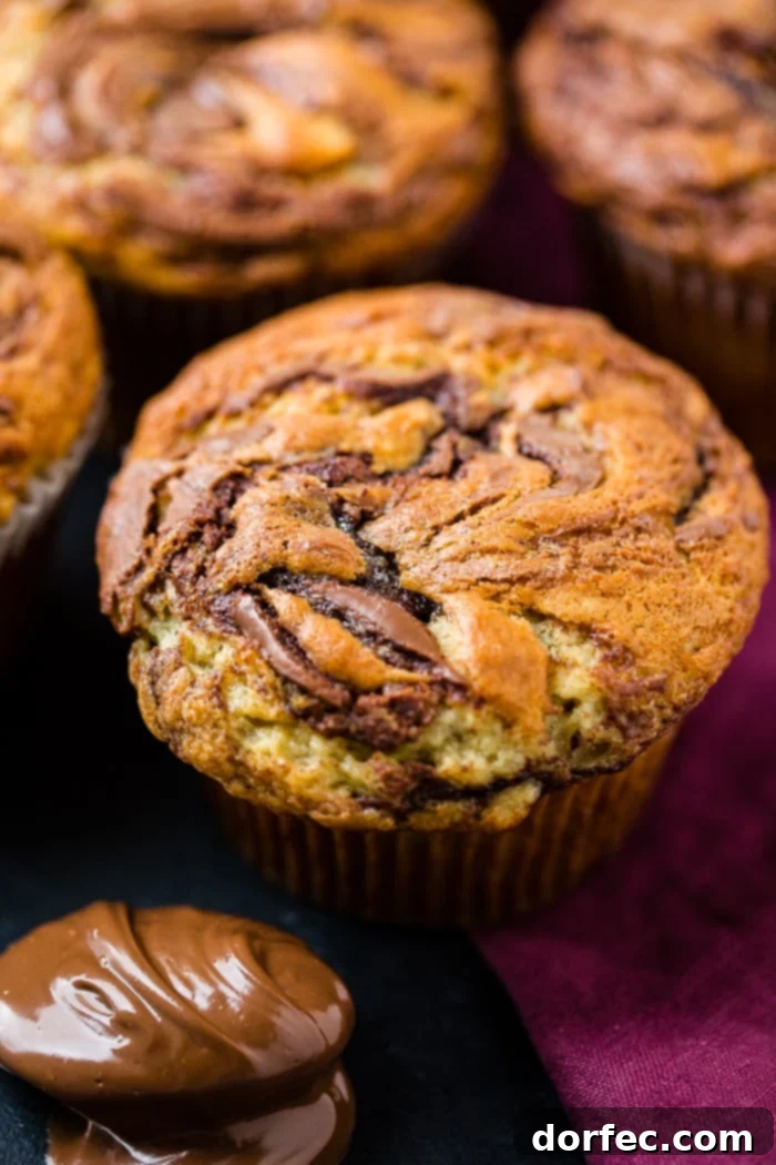 A close-up photograph of a moist Nutella swirled banana muffin with a golden-brown top, with more delicious muffins blurred in the background, enticing viewers to bake.