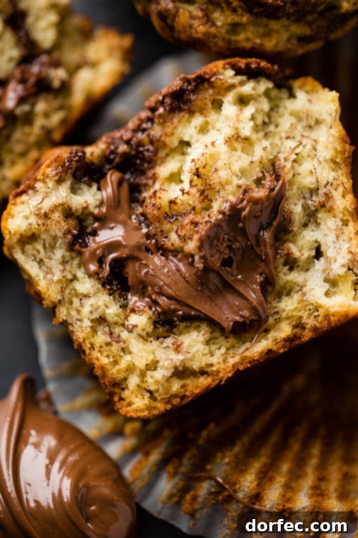 A perfectly baked, soft banana muffin cut in half, revealing a generous, gooey dollop of creamy Nutella spilling from its center, with more delicious muffins in the background.