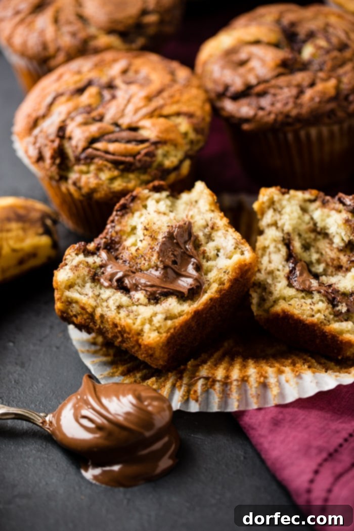 A collection of golden-brown Nutella swirled banana muffins, with one muffin cut open to reveal the rich, melted Nutella center, and a spoon with Nutella in the foreground.