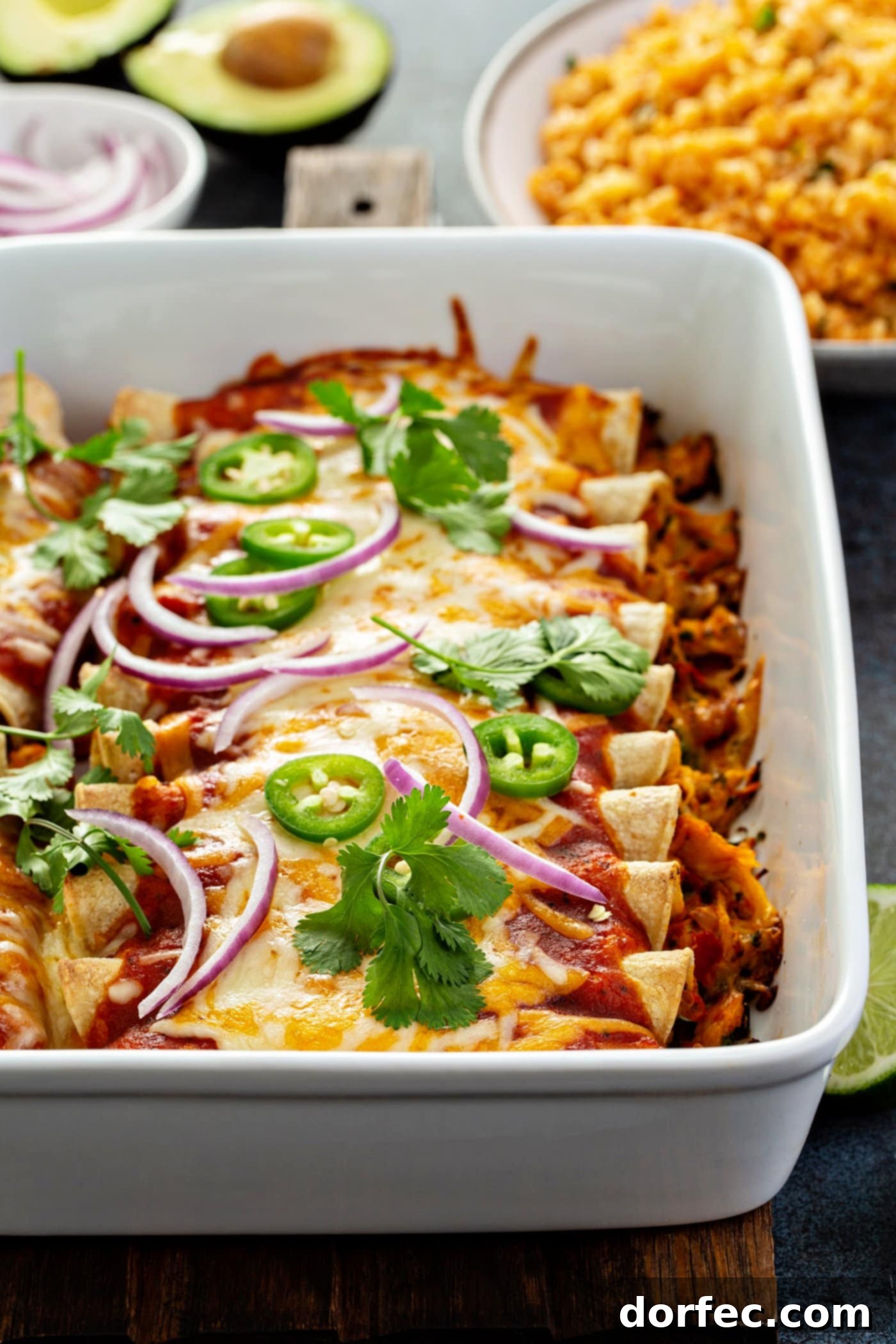 Red Chicken Enchiladas A white baking dish filled with baked red chicken enchiladas. They are topped with red sauce, melted cheese, cilantro leaves, slices of jalapeno and red onion.