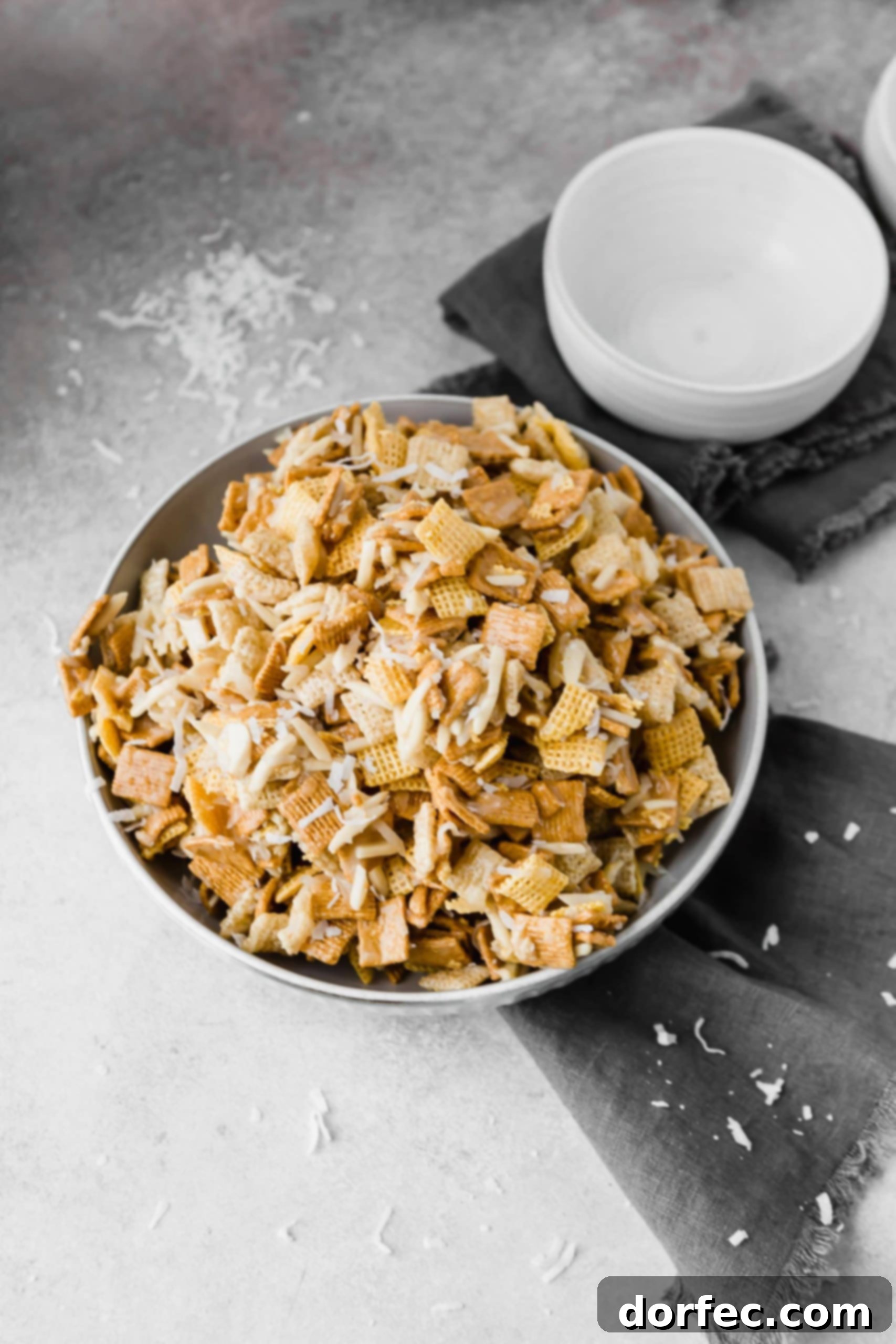 A large bowl brimming with golden-brown sweet gooey Chex mix, featuring Corn Chex, Rice Chex, Golden Grahams, slivered almonds, and shredded coconut, all coated in a shiny, sweet syrup.