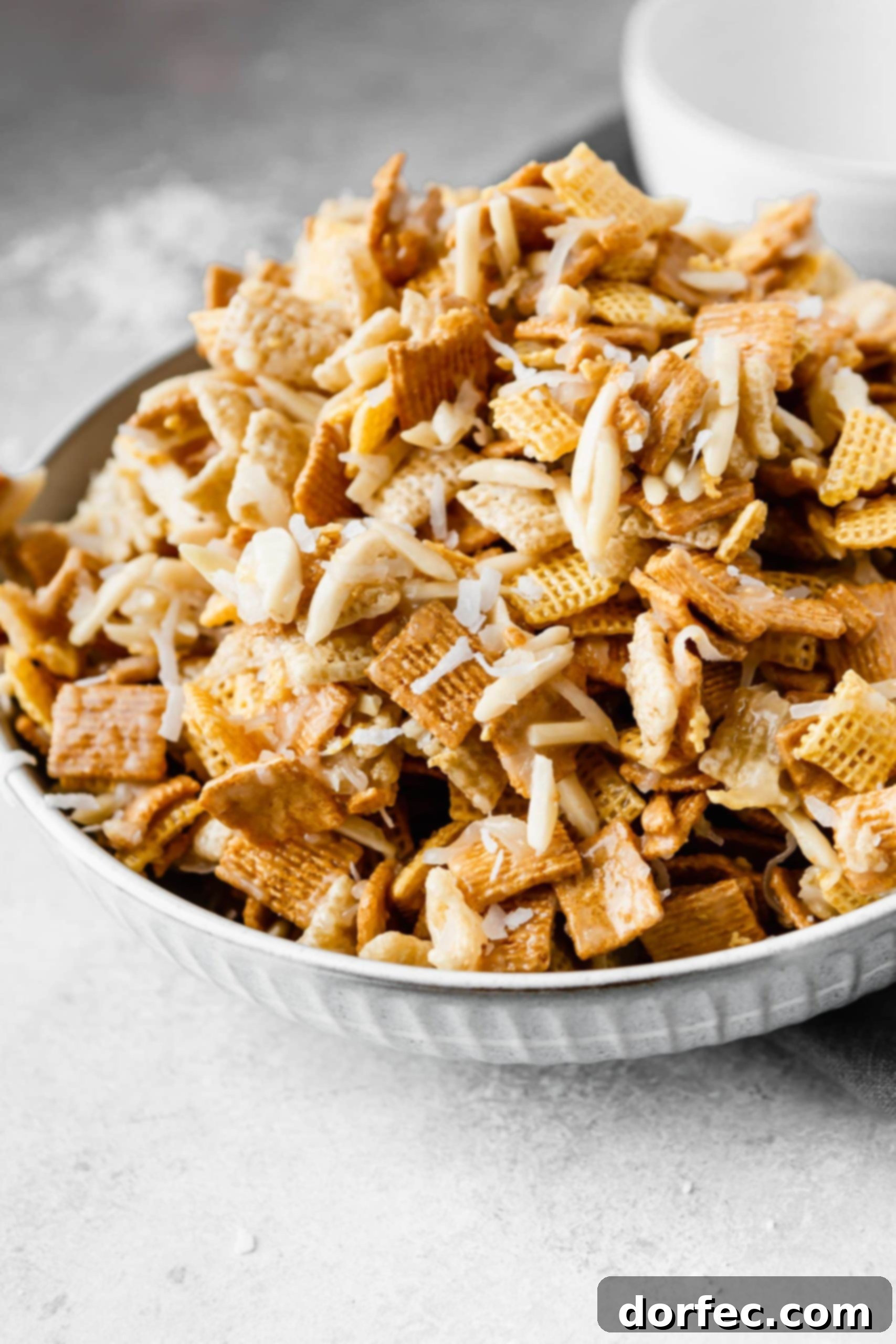 A large ceramic bowl overflowing with Sweet Chex Mix, filled with an assortment of Rice Chex, Corn Chex, Golden Grahams, slivered almonds, and glistening shredded coconut, all bound together by a luscious, sweet, and sticky coating.