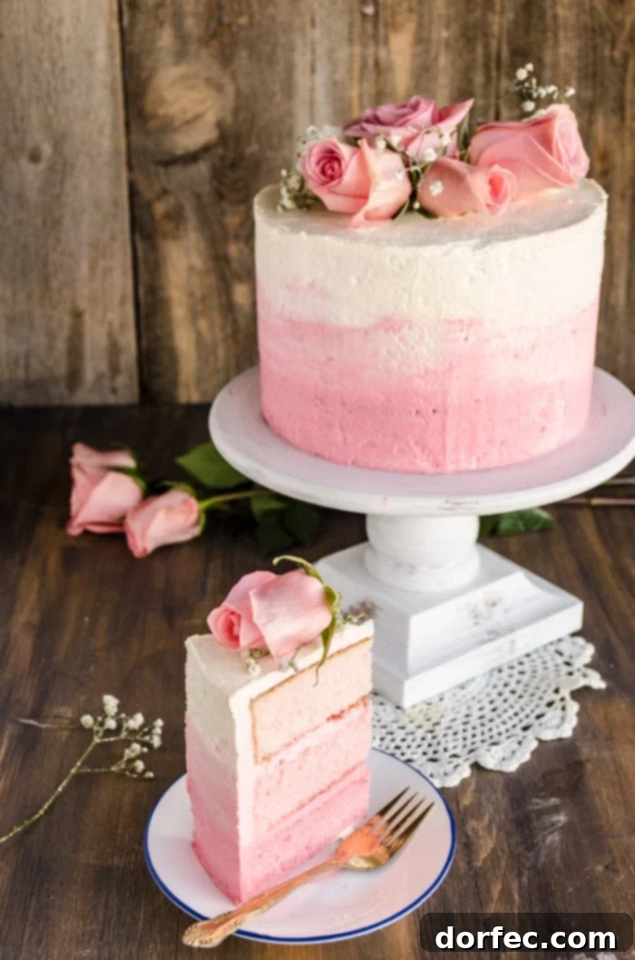 A stunning three-layer pink ombre cake topped with fresh roses and delicate sprigs of baby's breath, elegantly rests on a raised cake platter. Beside it, a perfectly cut slice of the ombre cake reveals its beautiful gradient of pink layers, inviting a delightful indulgence.