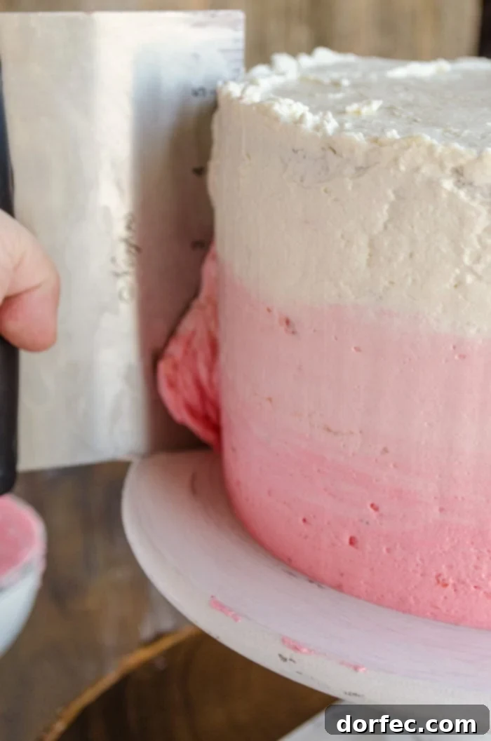 A three-layer Mother's Day Strawberry cake is topped with three shades of pink frosting. A hand holding a bench scraper is smoothing the sides of the cake, demonstrating the technique for a perfect ombre finish.