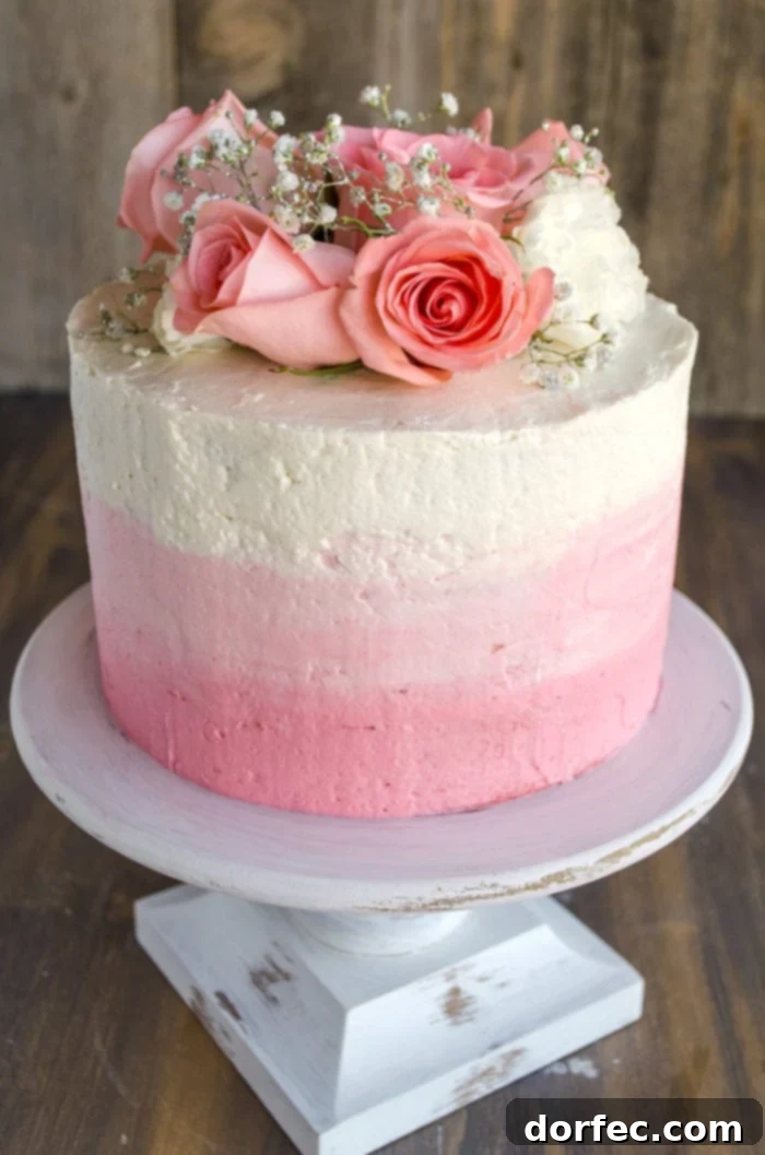 A stunning three-layer ombre Mothers Day strawberry cake, adorned with fresh pink roses and sparkling sprigs of baby's breath sitting atop a rustic white raised cake dish.
