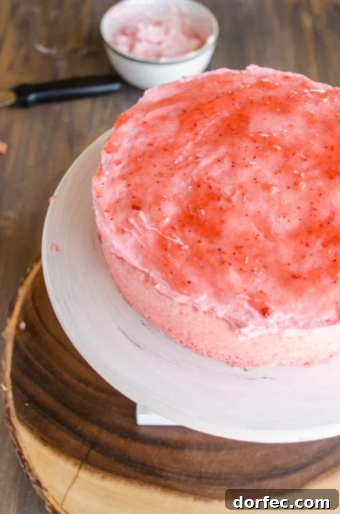 The deepest pink layer of the cake is positioned on a raised cake stand, with a corresponding pink frosting generously applied. A vibrant layer of strawberry jam crowns the frosting, adding flavor and color. In the background, a bowl of more pink frosting awaits its turn.