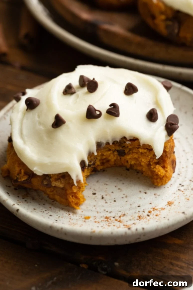 A thick, pillowy pumpkin chocolate chip cookie topped with swirls of cream cheese frosting and a sprinkle of mini chocolate chips, set against a cozy fall backdrop.