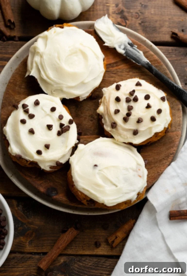 A wooden serving tray holds four luscious pumpkin chocolate chip cookies, each generously frosted with cream cheese frosting. Two are garnished with mini chocolate chips.