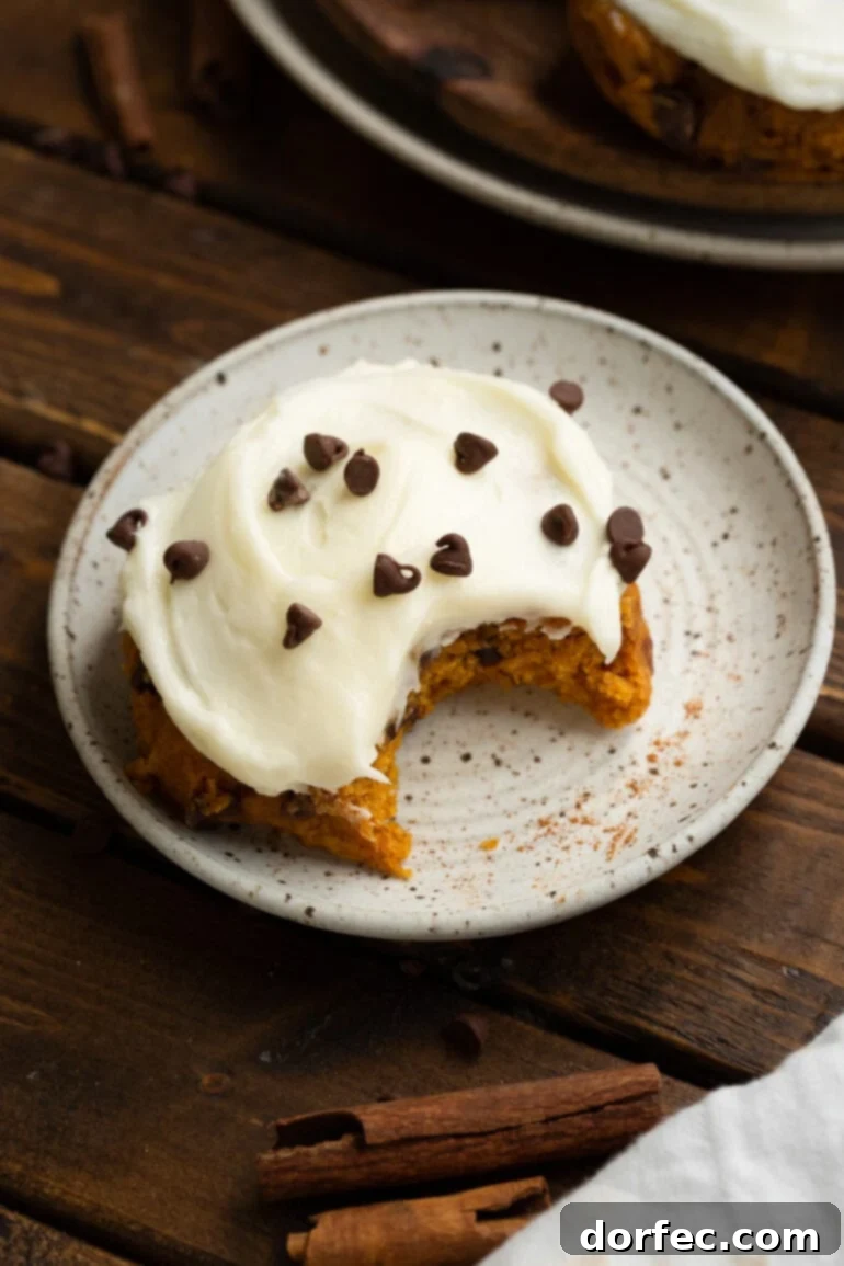 A frosted pumpkin chocolate chip cookie, garnished with mini chocolate chips, sits with a bite taken out, revealing its soft, cake-like interior.